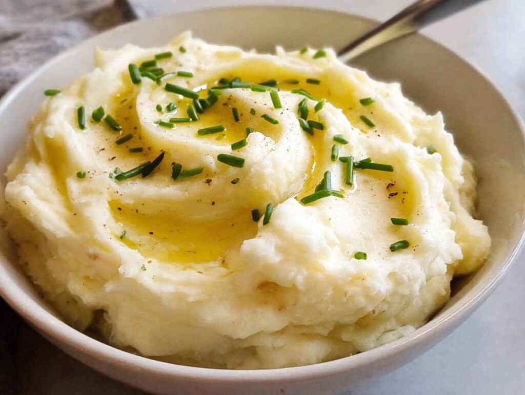A bowl of fluffy 12-Ingredient Mashed Potatoes topped with melted butter and fresh chives.