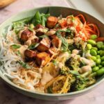 A vibrant and flavorful 20-ingredient rice bowl featuring crispy tofu, broccoli, edamame, shredded carrots, and noodles, drizzled with a creamy sauce.