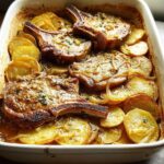 Close-up of baked pork chops with sliced potatoes in a casserole dish, featuring a rich sauce and herbs.