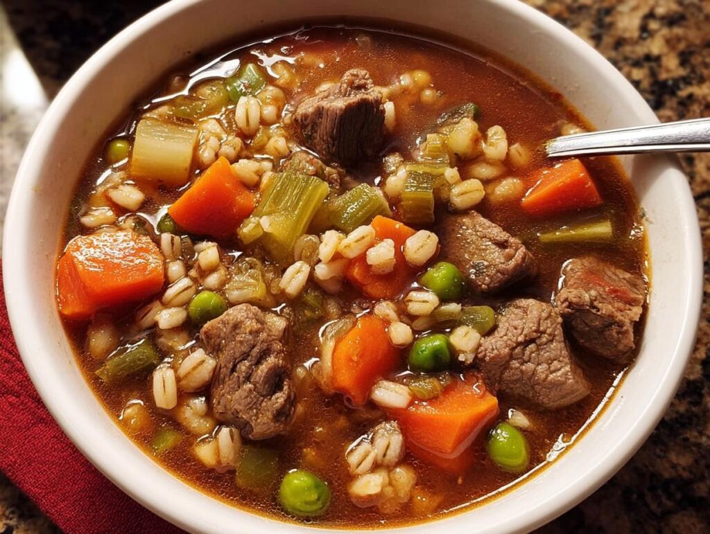 A close-up of a bowl of hearty beef barley soup, packed with tender beef chunks, carrots, peas, and barley.