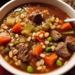 A close-up of a bowl of hearty beef barley soup, packed with tender beef chunks, carrots, peas, and barley.
