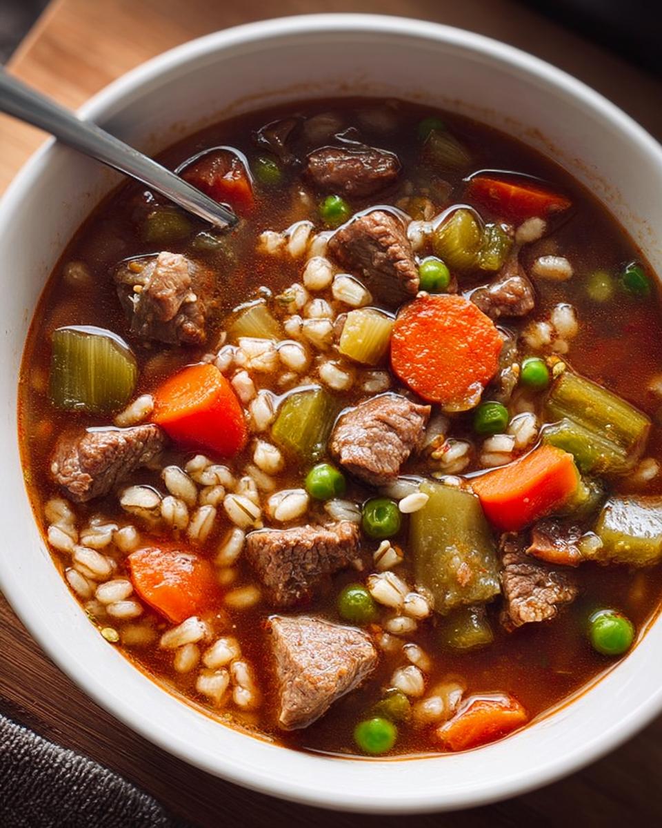 A close-up of a bowl of hearty beef barley soup, featuring tender chunks of beef, barley, carrots, celery, and peas in a rich broth.