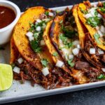 Close-up of three birria tacos with shredded beef, melted cheese, and cilantro, served with lime wedges and consommé for Taco Tuesday.