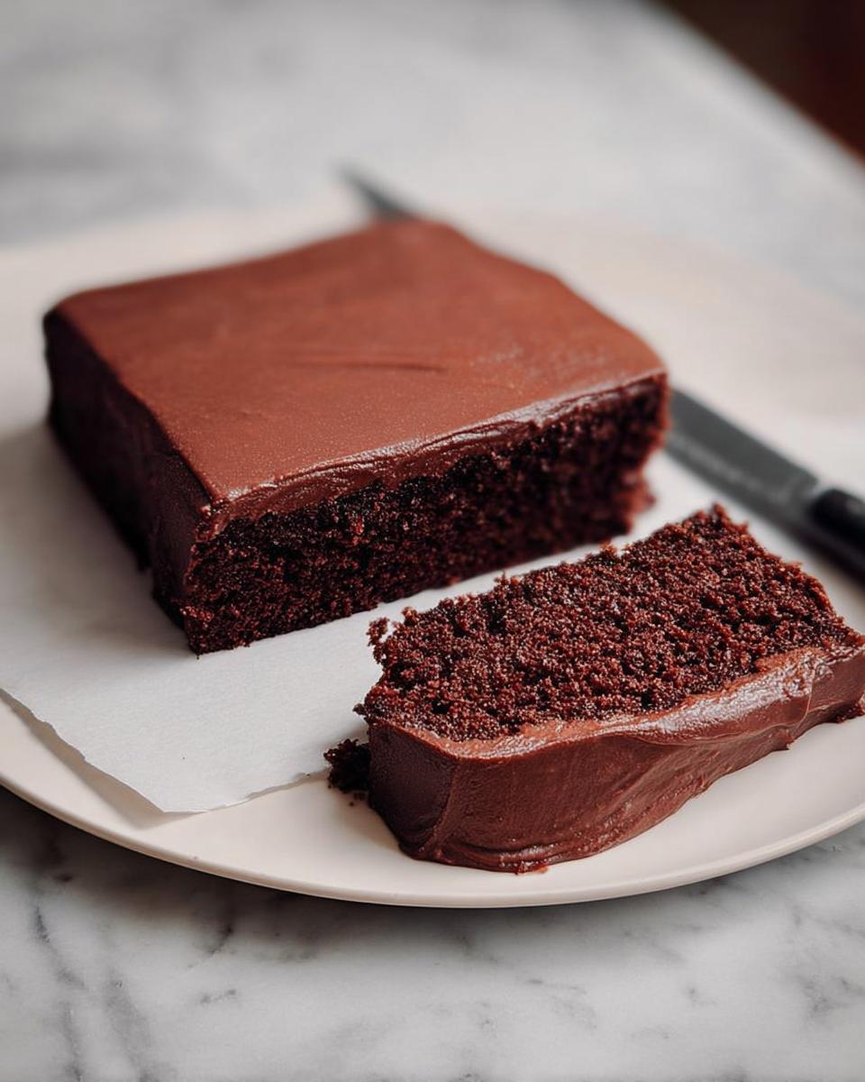 A slice of rich chocolate cake with smooth frosting, part of a larger cake on a plate.