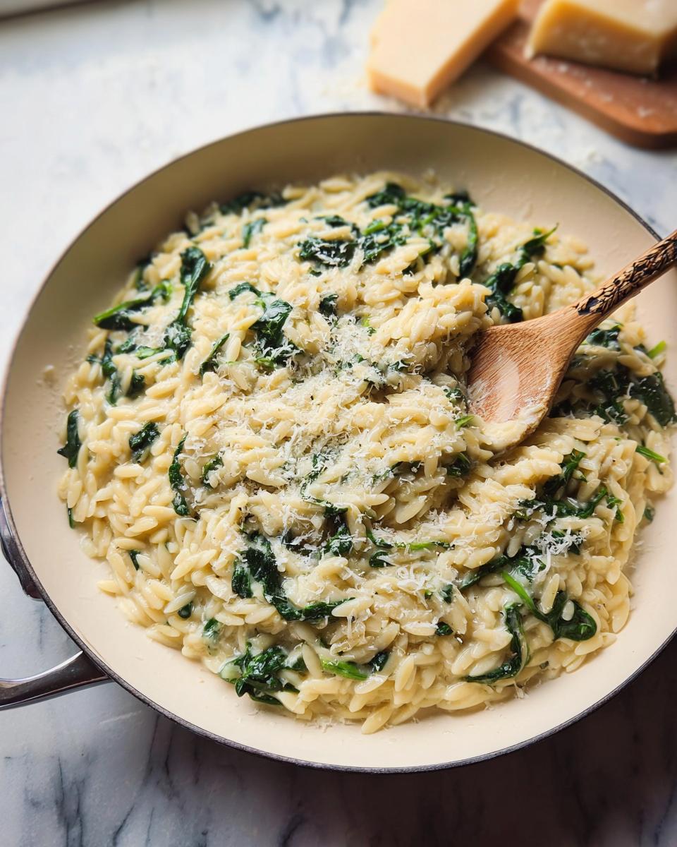 Creamy spinach orzo pasta dish in a pan, topped with grated parmesan cheese. A wooden spoon is mixing the dish.