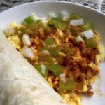 A close-up of a breakfast burrito next to a bowl of scrambled eggs with diced green peppers, onions, and a sprinkle of paprika.