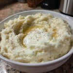 A bowl of creamy mashed potatoes topped with butter, herbs, and cracked black pepper.