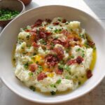 A bowl of creamy mashed potatoes topped with crispy bacon bits, fresh chives, and melted butter. A side of chopped chives is in the background.