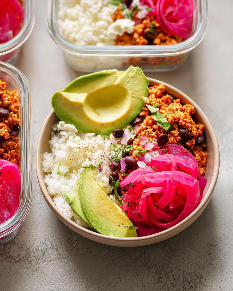 A colorful and healthy meal prep rice bowl featuring white rice, seasoned ground meat, black beans, avocado slices, and vibrant pickled red onions.