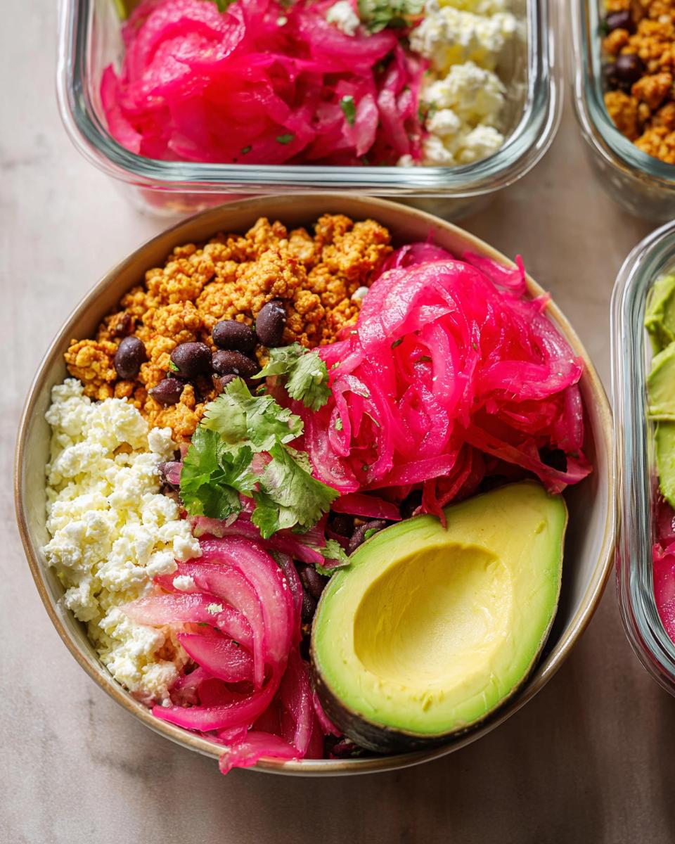 Close-up of a meal prep rice bowl with crumbled tofu, black beans, pickled red onions, avocado, and cheese.