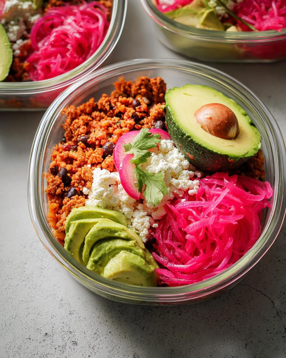 Close-up of a meal prep container filled with rice bowls recipes, featuring seasoned ground meat, black beans, avocado, feta cheese, and pickled red onions.