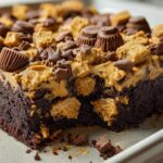 Close-up of a rich chocolate brownie cake topped with peanut butter frosting, chocolate chips, and peanut butter cup pieces.