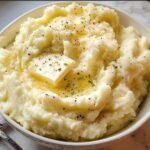 A bowl of fluffy mashed potatoes topped with a pat of butter and sprinkled with herbs and pepper.