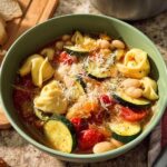 A bowl of quick tortellini soup with zucchini, tomatoes, and beans, topped with grated cheese.
