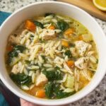 A close-up of a bowl of restaurant-style chicken orzo soup with carrots, spinach, and herbs.