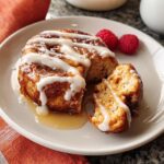 Close-up of a restaurant-style cinnamon roll drizzled with icing and syrup, served with raspberries.