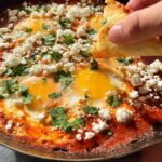 A hand dips bread into a skillet of restaurant-style eggs, shakshuka with feta cheese and parsley.