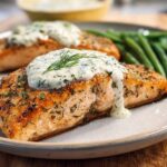 Close-up of a perfectly cooked salmon fillet topped with creamy dill sauce and fresh dill, served with green beans.