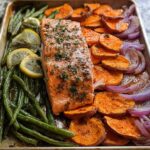 A delicious sheet pan meal prep featuring a salmon fillet with roasted sweet potatoes, green beans, red onions, and lemon slices.