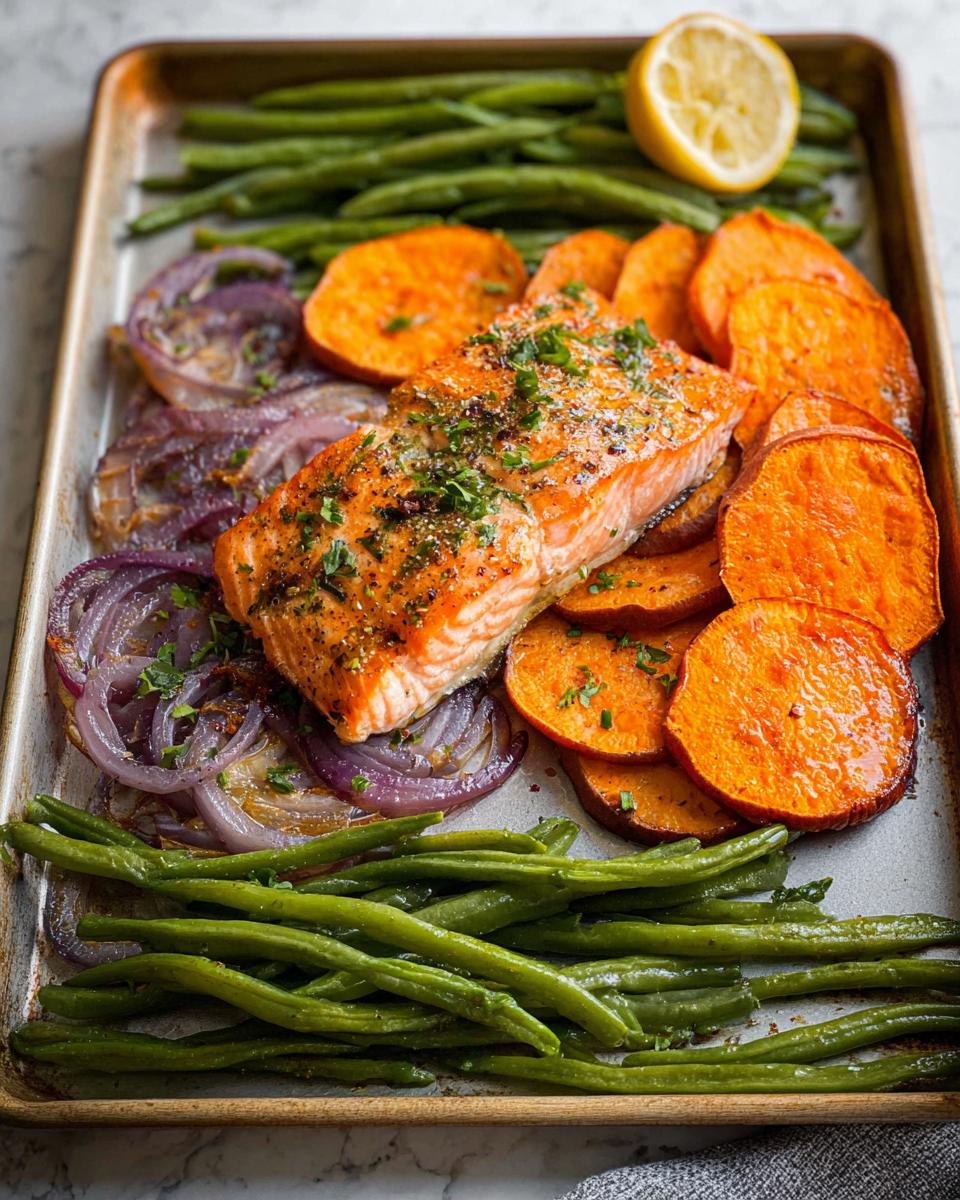 A perfectly cooked salmon fillet sits on a baking sheet with roasted sweet potatoes, red onions, and green beans, ideal for salmon recipes meal prep.