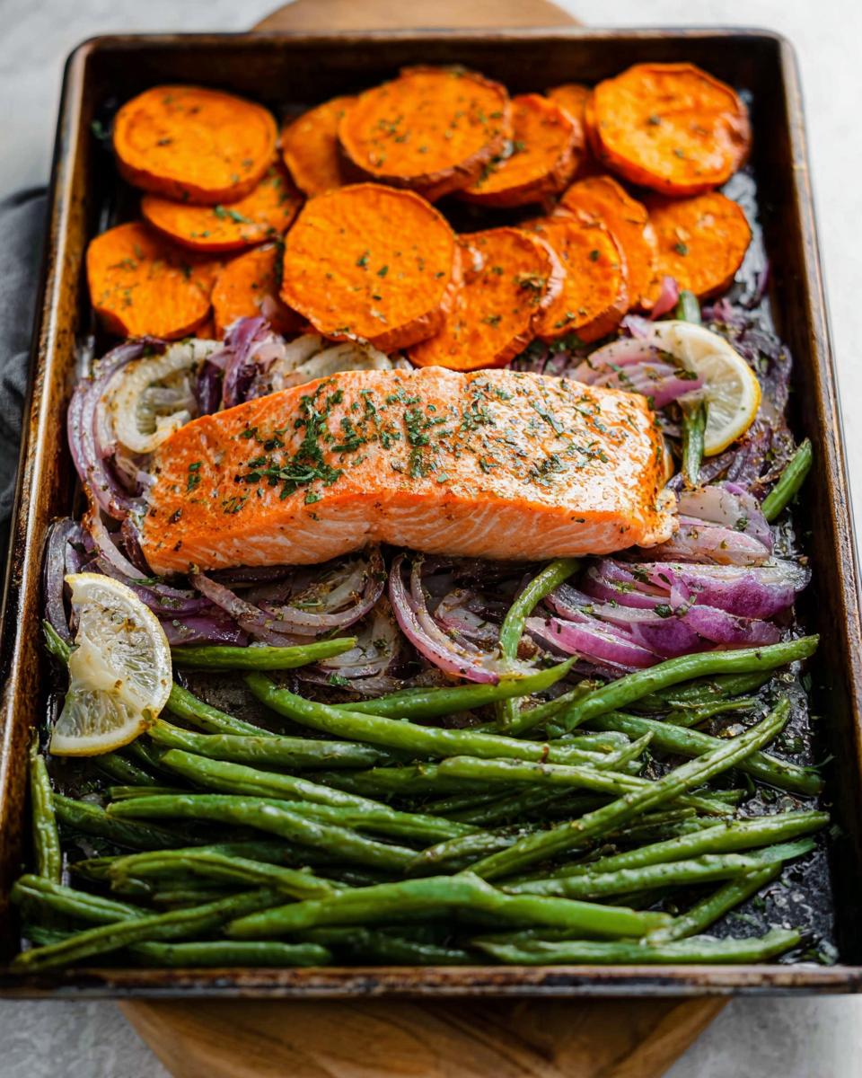 A delicious sheet pan meal prep featuring a salmon fillet, roasted sweet potato slices, green beans, and red onion.