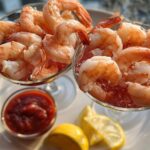 Two glasses filled with chilled shrimp cocktail, served with cocktail sauce and lemon wedges.