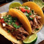 Two delicious shredded meat tacos for Taco Tuesday, topped with fresh cilantro, diced tomatoes, and onions, with lime wedges on the side.