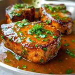 Close-up of three perfectly cooked pork chops swimming in a rich, glossy sauce and garnished with fresh parsley.