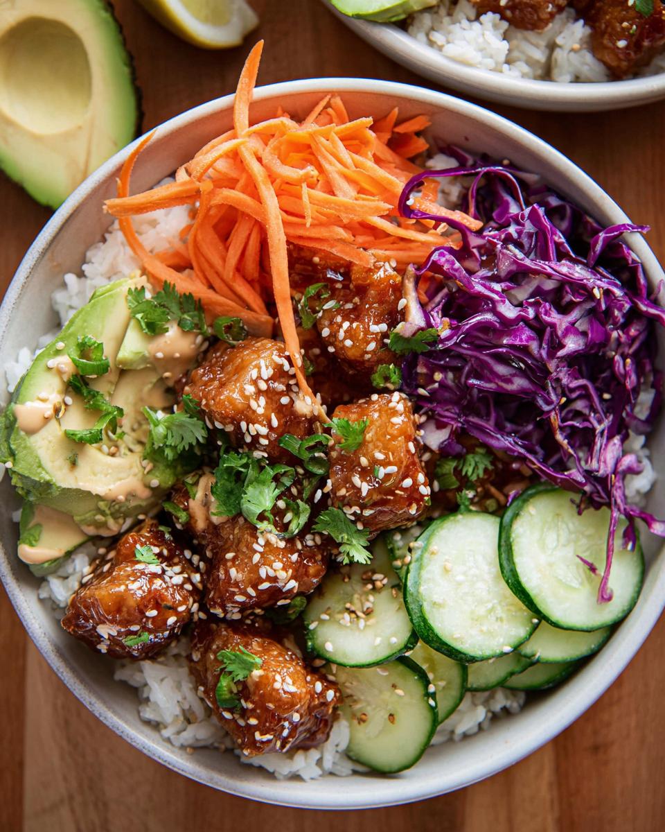 A close-up of a Bang Bang Chicken Bowl featuring crispy chicken, rice, avocado, shredded carrots, red cabbage, and cucumber slices.