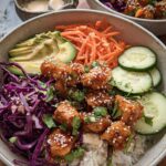 A vibrant Bang Bang Chicken Bowl filled with rice, crispy chicken, avocado, shredded carrots, red cabbage, and cucumber slices. Garnished with sesame seeds and cilantro.