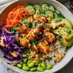 A vibrant bowl filled with rice, edamame, red cabbage, shredded carrots, cucumber slices, avocado, and topped with crispy Bang Bang Salmon Bites.