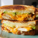 Close-up of a stacked CHICKEN AVOCADO MELT SANDWICH with melted cheese and fresh avocado slices.