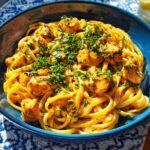 A close-up of Cowboy Butter Chicken Pasta in a blue bowl, featuring linguine noodles and tender chicken pieces in a creamy sauce, topped with fresh herbs.