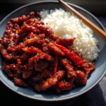 A close-up of a bowl of fluffy white rice next to glistening Crispy Chilli Beef, garnished with red chillies.