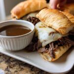 Close-up of a Crockpot French Dip Sandwich with melted cheese and a side of au jus.