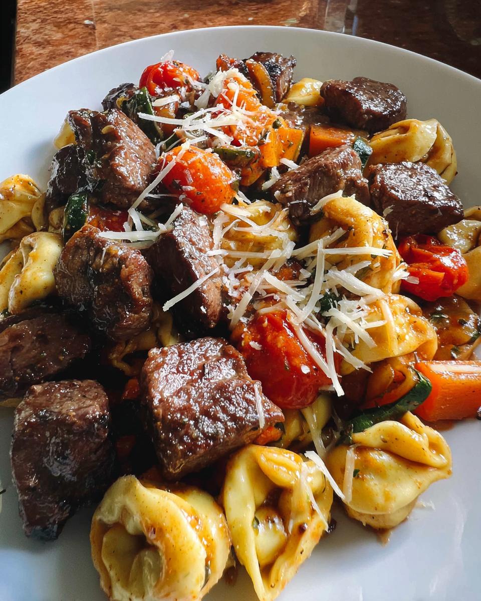 A close-up of a white bowl filled with Garlic Steak Tortellini, featuring tender steak bites, tortellini pasta, cherry tomatoes, and shredded cheese.