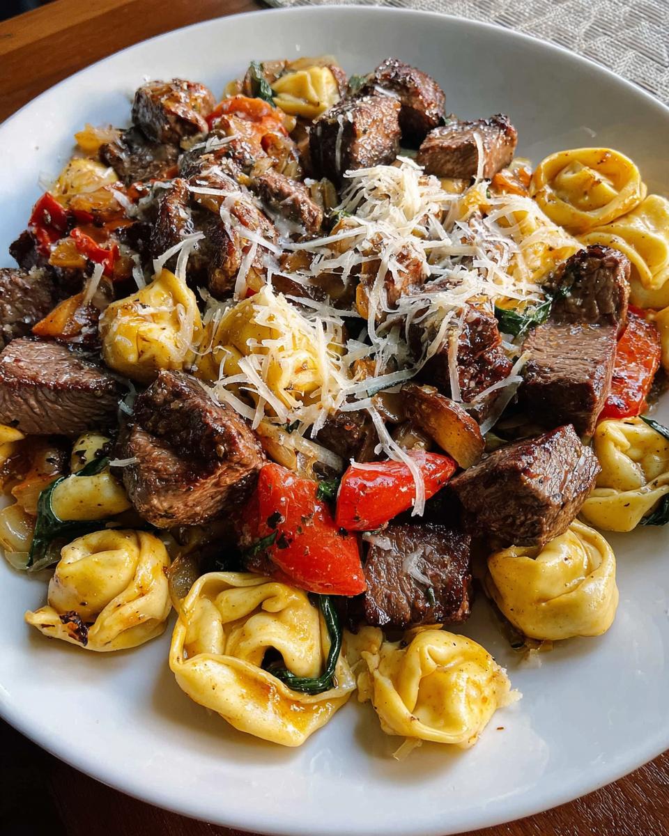 A close-up of a white plate filled with Garlic Steak Tortellini, featuring tender steak pieces, tortellini pasta, red peppers, and spinach, topped with grated cheese.