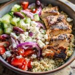 Close-up of a Greek Chicken Bowl with grilled chicken, quinoa, cucumber, tomatoes, red onion, feta cheese, and olives.