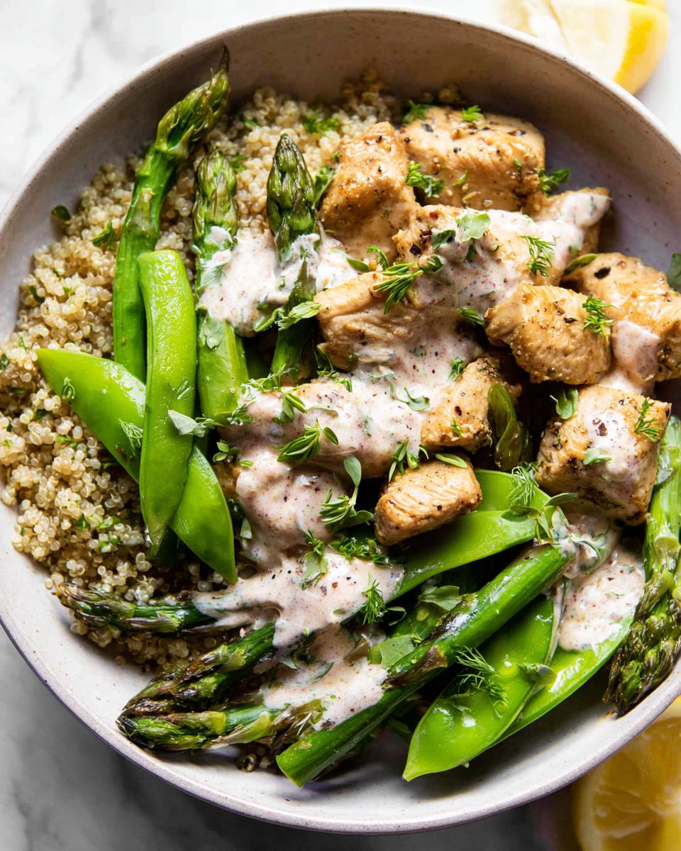 A healthy lemon garlic chicken meal prep bowl with quinoa, asparagus, and snap peas, drizzled with creamy sauce.