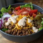 A delicious High-Protein Cheeseburger Bowl filled with seasoned ground beef, shredded cheese, lettuce, tomatoes, onions, and pickles, drizzled with sauce.