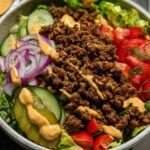 A delicious High-Protein Cheeseburger Bowl filled with seasoned ground beef, lettuce, tomatoes, cucumbers, red onion, and pickles, drizzled with sauce.