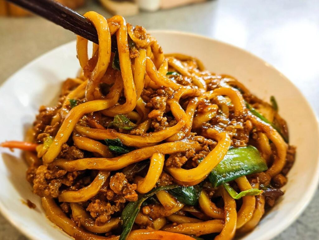 Close-up of a bowl of Irresistible Yaki Udon noodles with ground meat and vegetables, being lifted with chopsticks.