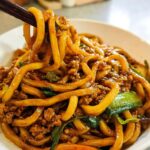 Close-up of a bowl of Irresistible Yaki Udon noodles with ground meat and vegetables, being lifted with chopsticks.