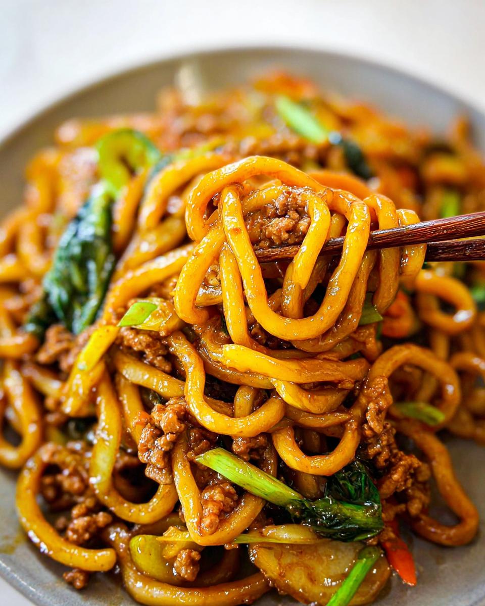 Close-up of a plate of Irresistible Yaki Udon Noodles with ground meat and green vegetables, being lifted with chopsticks.