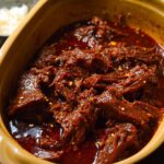Close-up of a rich, dark sauce covering tender shredded Korean Style Pot Roast in a yellow ceramic dish.