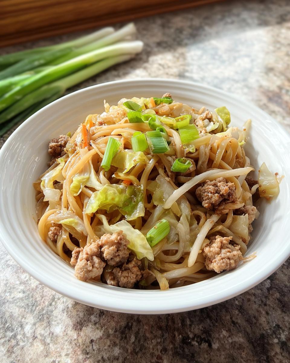 A close-up of a white bowl filled with a Potsticker Noodle Bowl featuring noodles, ground pork, cabbage, and green onions.