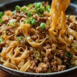A close-up of a steaming Potsticker Noodle Bowl with Pork & Cabbage Slaw, featuring wide noodles, ground pork, and green onions.
