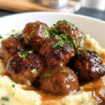 A close-up of Salisbury steak meatballs covered in gravy, served over garlic herb mashed potatoes and garnished with parsley.