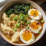 A close-up of a Savory Dumpling Ramen Bowl with soft-boiled eggs, noodles, dumplings, and greens.