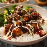 A close-up of a bowl filled with white rice, topped with glazed sticky chicken pieces and steamed broccoli florets, drizzled with sauce and sesame seeds.